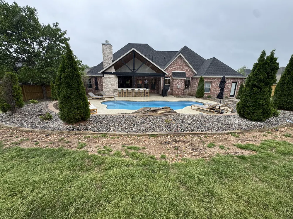 Landscaped beds and shaped evergreens wrapping a pool behind a large brick home