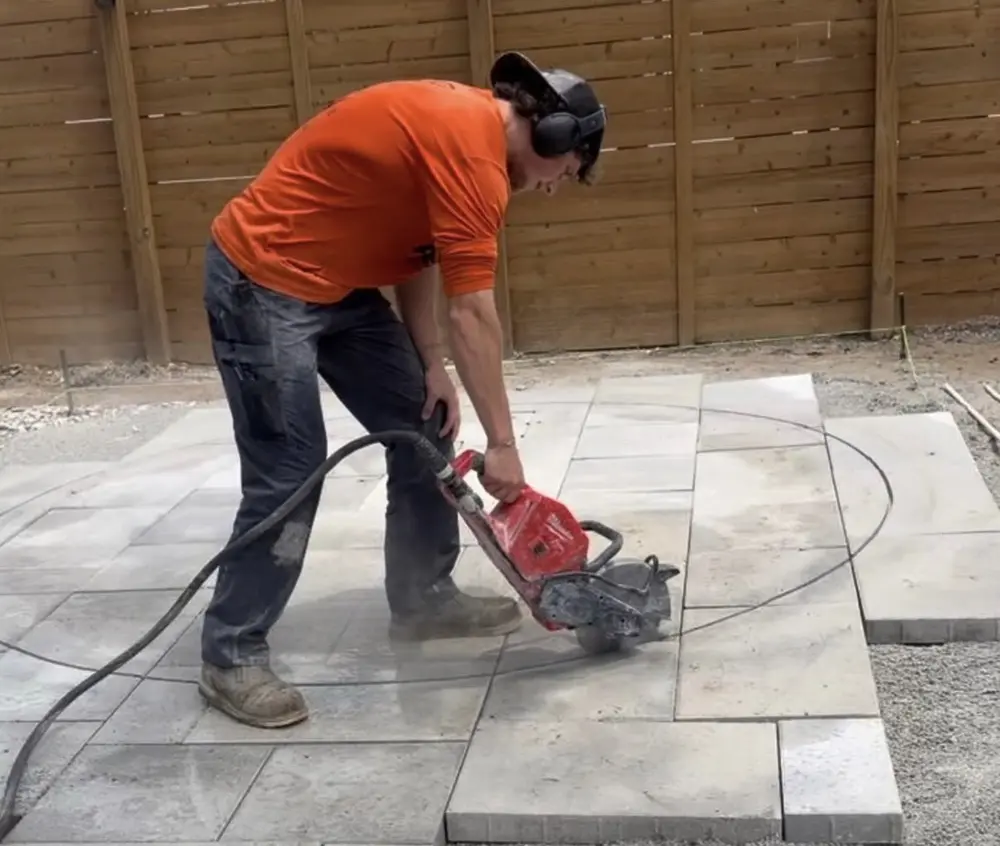 Peak installer in an orange shirt cutting a paver with a cut off saw on a new patio