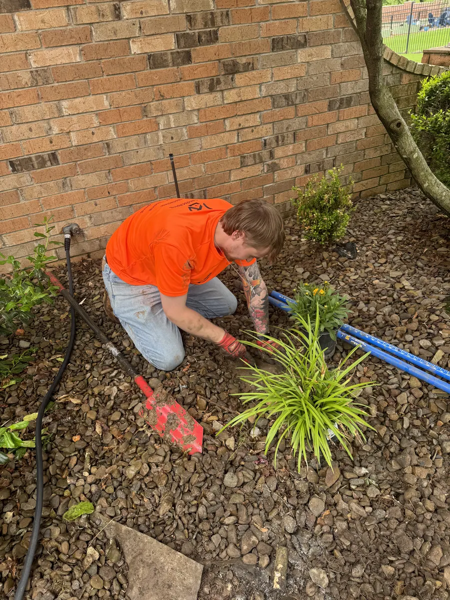 Peak crew member in an orange shirt setting a plant into a river rock bed against a brick wall