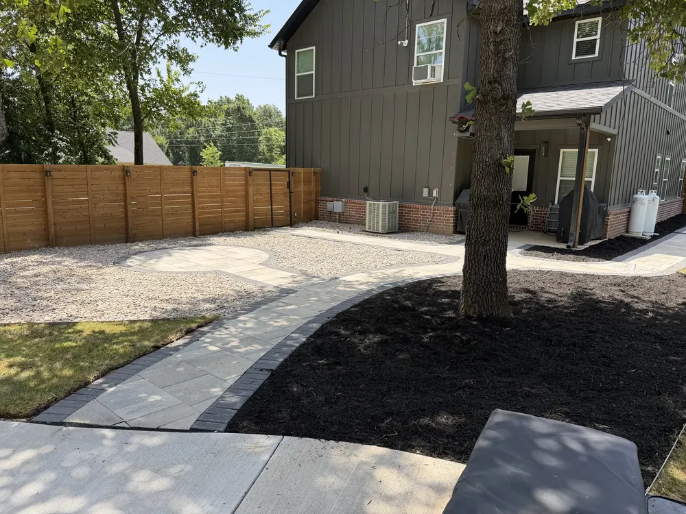 Paver walk running past a mature tree set in a black mulch bed inside a fenced back yard